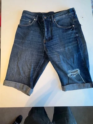 Blå denimshorts från &Denim - Säljer ett par blå denimshorts från &Denim i rak passform. Shortsen har klassisk femficksdesign, uppvikta benslut och en snygg sliten detalj på ena benet. Perfekta för en avslappnad stil under varmare dagar.