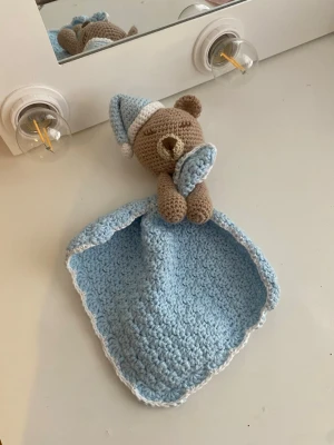 Virkad snuttefilt med nalle - Handvirkad snuttefilt i ljusblått bomullsgarn med en brun nallebjörn som har en matchande blå sovmössa. 