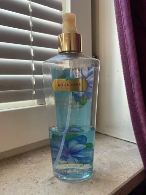 Victoria's Secret Aqua Kiss mist - Fräsch body mist från Victoria's Secret i doften Aqua Kiss. Flaskan är genomskinlig med blått innehåll och dekorerad med blå blommor samt gulddetaljer på etikett och sprayhuvud. Perfekt för dig som gillar lätta, blommiga dofter.