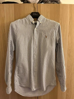 Randig skjorta från Ralph Lauren - Klassisk vit och blå randig skjorta från Ralph Lauren med button-down krage och broderad logga på bröstet. Skjortan har långa ärmar och knäppning framtill. Perfekt på sommaren med svalkande materialet och passformen!
