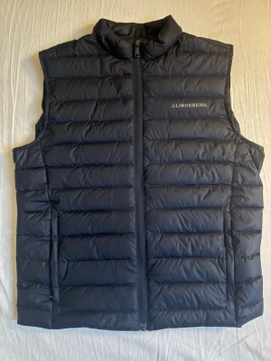 Svart dunväst från J.Lindeberg - Snygg svart dunväst från J.Lindeberg med quiltad design och diskret logga på bröstet. Västen har hög krage och dragkedja framtill, perfekt att slänga över en hoodie eller t-shirt för en clean look.