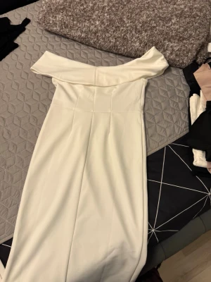 Vit offshoulder långklänning - Säljer en elegant vit långklänning med offshoulder-design. Klänningen har en figurnära passform och stilren look, perfekt för speciella tillfällen. Materialet känns mjukt och följsamt, troligtvis polyester eller liknande tyg.