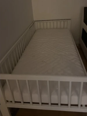 Vit barnsäng med ribbor - Säljer en vit barnsäng med ribbade sidor och huvudgavel. Sängen har en enkel, modern design och är tillverkad i trä. Madrass ingår och sängen passar perfekt till barnrummet.