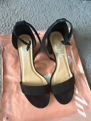 Svarta chunky klackpumps från SheIn - Säljer ett par svarta pumps från SheIn med chunky klack och öppen tå. Skorna har en bred rem över tårna och justerbart ankelband med spänne. Materialet är matt tyg och innersulan är beige. Perfekta för att lyfta vilken outfit som helst.