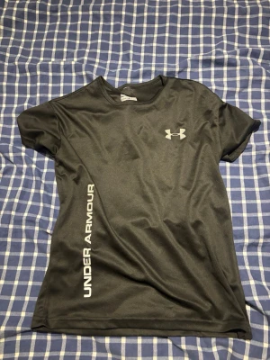 Svart träningströja från Under Armour - Snygg svart träningströja från Under Armour med korta ärmar och rund hals. Tillverkad i lätt och snabbtorkande polyester, perfekt för intensiva träningspass. Logotyp på bröstet och text längs sidan ger en sportig look.