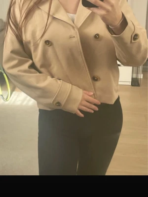 Kort beige dubbelknäppt rock från lindex - Snygg kort beige rock med dubbelknäppning och stora bruna knappar. Rocken har bred krage och långa ärmar med knappdetaljer vid ärmsluten. Perfekt för dig som gillar en stilren och trendig look. Materialet är mjukt och bekvämt. Nyskuck aldrig använd!!