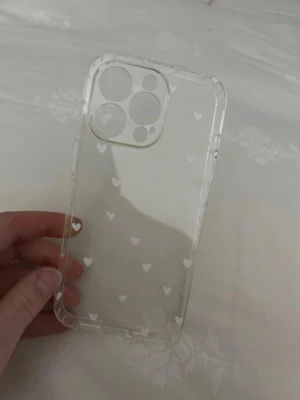 Genomskinligt mobilskal med vita hjärtan för iPhone 13Pro  - Säljer ett genomskinligt mobilskal med små vita hjärtan. Skalets kanter är förstärkta för extra skydd och det är gjort i mjuk plast som känns skön att hålla i. Passar till iPhone 13 Pro. Jag beställde fel så den är helt ny! 