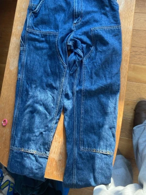 Carhartt jeans - Säljer ett par blå jeans med raka ben och stora, praktiska fickor både fram och bak. Jeansen har tydliga sömmar och är tillverkade i klassiskt denimtyg. Perfekta för en avslappnad och cool stil. Storlek: 27 32