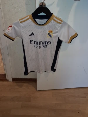 Real Madrid matchtröja Bellingham - Säljer en vit Real Madrid matchtröja från Adidas med guldiga detaljer och svarta inslag. Tröjan har klubbmärke, sponsortryck och Bellingham 5 på ryggen. Perfekt för dig som älskar fotboll och vill stötta ditt lag på match eller träning.
