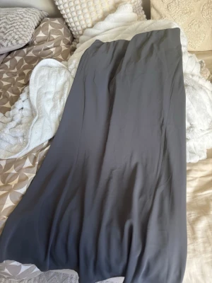 Mörkgrå lång satinkjol - Säljer en stilren långkjol i mörkgrått satinliknande material. Kjolen har en mjuk, glansig yta och faller snyggt längs kroppen. Perfekt för dig som gillar minimalistisk och clean stil.