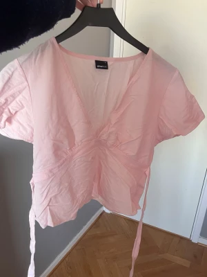 Ljusrosa omlottopp från Gina Tricot - Supersöt ljusrosa omlottopp från Gina Tricot med korta ärmar och knytband i midjan. Har en liten fläck men den går säkert bort i tvätten, annars bra skick🩷passar XS-M eftersom man kan justera snöret beroende på passform.