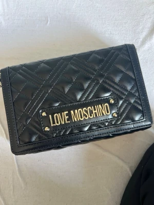 Svart quiltad handväska Love Moschino - Snygg svart handväska från Love Moschino i quiltat konstläder med guldfärgad logotyp framtill. Väskan har rektangulär form och stilrena detaljer som ger en lyxig känsla. Perfekt accessoar för att lyfta din outfit.