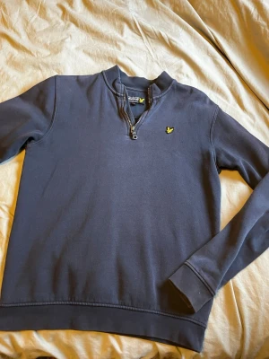 Mörkblå Lyle & Scott halvzip tröja - Mörkblå långärmad tröja från Lyle & Scott med halvzip och gul logga på bröstet. Tröjan har ribbade muddar och en mjuk bomullskänsla, perfekt för en avslappnad och stilren look.
