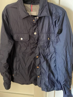 Mörkblå vindjacka från Moncler - Snygg mörkblå vindjacka från Moncler med klassisk krage, två bröstfickor med lock och tryckknappar samt svart Moncler-logga på ärmen. Jackan är tillverkad i ett lätt, glansigt material som står emot vind. Perfekt för dig som vill ha en stilren och sportig look.