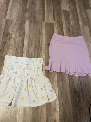 Två korta kjolar med volang - Säljer två snygga kortkjolar. En vit kjol med gult blommönster och smock i midjan, samt en ljuslila kjol med volangkant nertill. Perfekta för sommarens alla tillfällen!