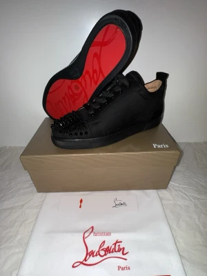 Svarta Christian Louboutin sneakers - Svarta låga sneakers från Christian Louboutin med mattsvarta nitar på tån och klassisk röd sula. Skorna har snörning och är tillverkade i mocka och läder. Perfekta för dig som vill sticka ut med en edgy och lyxig stil.