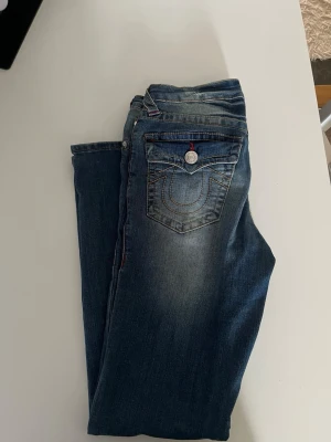Blå True Religion jeans med kontrastsömmar - Säljer ett par blå jeans från True Religion med ikonisk kontrastsöm och snyggt slitna detaljer. Jeansen har klassisk femficksmodell och knappstängning. Bakfickan har den typiska hästsko-loggan och silverfärgad knapp. Perfekta för en avslappnad och trendig look. Jeansen är i storlek W25 L29 och sitter någon centimeter för långt på mig, som är 153 cm. De är inte helt lowrise, men sitter ändå någorlunda lågt. Mycket bra skick och knappt använda.