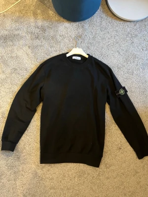 Svart sweatshirt från Stone Island - Snygg svart sweatshirt från Stone Island med klassisk rund halsringning och ikonisk logotyp-patch på vänster ärm. Tröjan har ribbade muddar vid ärmslut och nederkant. Perfekt för en clean och stilren look.