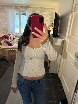 Vit blommig långärmad topp - Söt tunn långärmad topp med små blommor i ljusblått. Toppen har en croppad passform, rundad ringning och knappar framtill. Perfekt att styla med jeans eller kjol för en chill look.