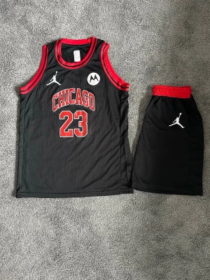 Chicago Bulls basketset Jordan #23 - Snyggt basketset med svart linne och shorts från Jordan, inspirerat av Chicago Bulls. Linnet har röda detaljer, texten 'CHICAGO' och nummer 23 framtill. Både linne och shorts har Jumpman-loggan. Tillverkat i lätt och ventilerande material, perfekt för basket eller träning.