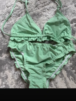 Grön bikini med volangdetalj - Snygg ljusgrön bikini med volangdetalj på överdelen och klassiska bikinitrosor. Överdelen har trekantskupor och knyt i nacken. Perfekt för lata dagar på stranden eller poolen.