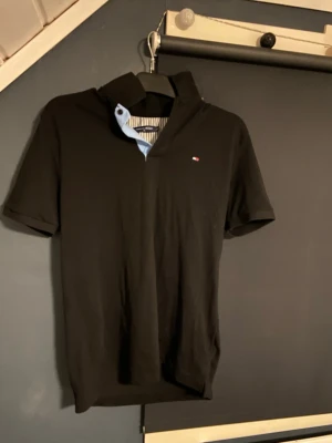 Svart piké från Tommy Hilfiger - Snygg svart pikétröja från Tommy Hilfiger med liten logga på bröstet och klassisk krage med blå insida. Perfekt till jeans eller shorts och har en stilren look. Tillverkad i mjuk bomull och har korta ärmar.