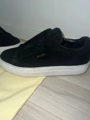 Svarta sneakers från Axel Arigato - Stilrena svarta sneakers från Axel Arigato i mocka med diskret guldfärgad logga på sidan. Skorna har en chunky vit sula och minimalistisk design utan synlig snörning. Perfekta för dig som gillar clean och modern stil.