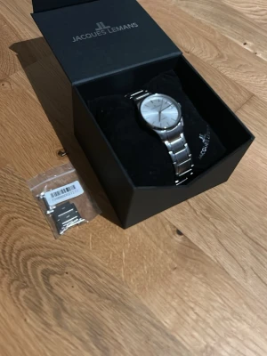 Jacques Lemans silverfärgad klocka - Stilren klocka från Jacques Lemans i silverfärgat rostfritt stål med klassisk länkarmband och rund urtavla. Levereras i originalask och extra länkar medföljer för justering av storlek. Perfekt accessoar för en modern look.