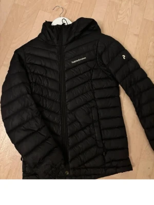 Svart dunjacka Peak Performance XS - Snygg svart dunjacka från Peak Performance med huva och dragkedja framtill. Jackan har två sidofickor med dragkedja och diskret logga på bröstet samt ärmen. 