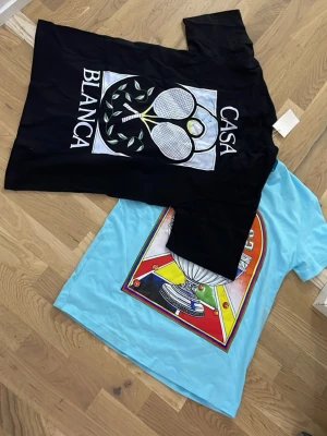Casablanca t-shirts med tryck - Två t-shirts från Casablanca, en svart med stort grafiskt tennistryck och texten 'Casa Blanca', och en ljusblå med färgglatt tryck och 'Casablanca Tennis Club'. Båda har rund hals och korta ärmar. Perfekta för dig som gillar unika prints och sportig stil.