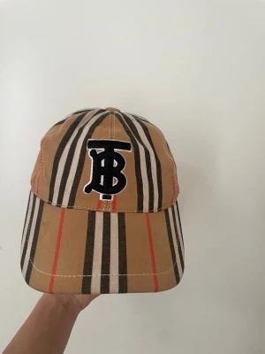 Randig keps från Burberry - Snygg keps från Burberry med klassiskt beige, svart, vitt och rött randigt mönster. Framtill finns ett broderat svart och vitt TB-monogram. Justerbar rem baktill. Perfekt accessoar för en trendig streetstyle-look.