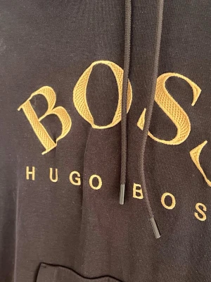 Svart hoodie från Hugo Boss - Snygg svart hoodie från Hugo Boss med stor gul broderad logga på bröstet. Klassisk känguruficka framtill, ribbade muddar och justerbar huva med snören. Perfekt för dig som vill ha en stilren och trendig look.