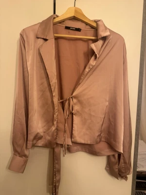 Rosa satinblus med knyt från BikBok - Snygg ljusrosa satinblus från BikBok med långa ärmar och knyt framtill. Blusen har klassisk krage och en glansig finish som ger en lyxig känsla. Perfekt att styla med jeans eller kjol för en trendig look.