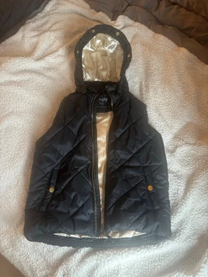 Svart quiltad dunväst med huva - Snygg svart dunväst från Only med quiltat mönster och guldfärgade knappar på fickorna. Västen har en avtagbar huva med glansigt beige innerfoder och dragkedja framtill. Perfekt för lager på lager och ger en cool look till din outfit.