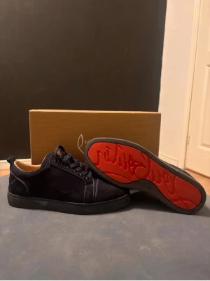 Mörkblå Louboutin sneakers med röd sula - Snygga mörkblå sneakers från Christian Louboutin i mocka med klassisk röd sula och svarta snören. Låg modell med stilren design och exklusiv känsla. Perfekta för dig som vill sticka ut med en ikonisk detalj på fötterna.