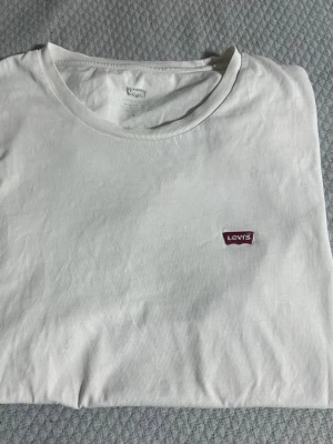 Vit t-shirt från Levi's med logga - Klassisk vit t-shirt från Levi's med rund halsringning och den ikoniska röda Levi's-loggan broderad på bröstet. Tillverkad i mjuk bomull för en skön och avslappnad stil. Perfekt basplagg till alla outfits.