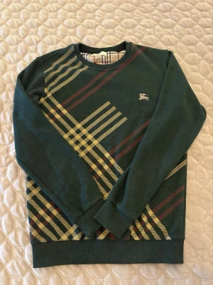 Mörk Grön stickad tröja från Burberry - Snygg grön stickad tröja från Burberry med klassiskt rutmönster i beige, rött och svart. Tröjan har rund halsringning, långa ärmar och ribbade muddar. Ikoniska Burberry-loggan broderad på bröstet. Perfekt för dig som gillar stilrena detaljer.