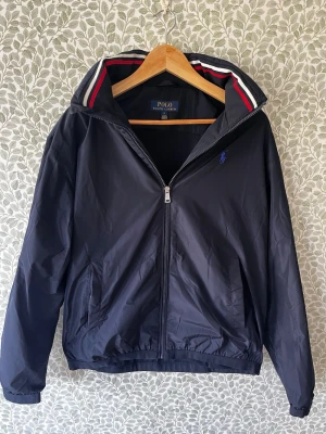 Svart jacka från Polo Ralph Lauren - Tjenare! Säljer nu min mörkblå regnjacka från Polo Ralph Lauren då den är något för stor. Jackar har dragkedja, inbyggd huva och elastiska muddar. Huvkanten är dekorerad med röda och vita ränder och den klassiska blåa broderade loggan sitter förstås på bröstet. Materialet är lätt och smidigt, perfekt för höst, vår och sommar. Har bara använts ett par gånger så skicket är klockrent, hör gärna av dig vid minsta fundering!