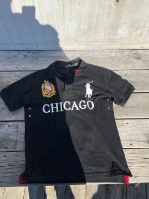 Svart pikétröja Polo Ralph Lauren - Svart pikétröja från Polo Ralph Lauren med broderad logga och texten 'CHICAGO' på bröstet. Tröjan har korta ärmar, krage och knappar framtill samt detaljer i vitt och gult broderi. Perfekt för en sportig och stilren look.