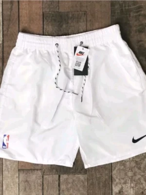 Vita Nike shorts med snörning NBA - Säljer ett par vita shorts från Nike med svart snörning i midjan och NBA-logga på benet. Shortsen har elastisk midja och är perfekta för sport eller chill. Klassisk Nike Swoosh på ena benet.