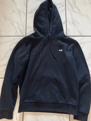 Svart hoodie från JJR med tryck - Svart hoodie från JJR med liten vit logga på bröstet. Klassisk modell med huva och dragsko, ribbade muddar vid ärmslut och nederkant. Perfekt för en chill och avslappnad stil. Materialet känns mjukt och bekvämt.