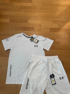 Vitt träningsset från Under Armour -  Vitt träningsset från Under Armour med t-shirt och shorts. Setet har svarta loggor och tryck, samt elastisk midja på shortsen
