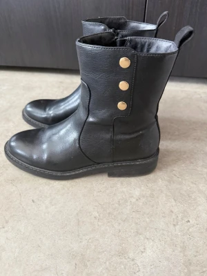 Svarta boots med gulddetaljer - Svarta boots i skinnimitation med rund tå och tre guldfärgade knappar på sidan. Dragkedja på insidan och ögla baktill för enkel påtagning. Klassisk och stilren design som passar till många outfits.