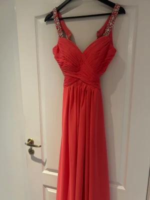 Korallfärgad långklänning med strass - Snygg korallfärgad långklänning med plisserad överdel och breda axelband dekorerade med strass. Klänningen har en öppen rygg med unika banddetaljer och ett flytande, elegant fall. Perfekt för speciella tillfällen. Skriv för flera bilder eller prisförslag 💞💞