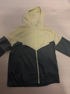 Nike tvåfärgad vindjacka med huva - Snygg vindjacka från Nike i tvåfärgad design med ljusgul ovandel och mörkgrå underdel. Jackan har hel dragkedja, huva och två sidofickor. Nike-logga på bröstet. Perfekt för blåsiga dagar och streetwear-stil.