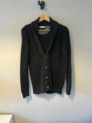 Mörkgrå stickad kofta med knappar - Mysig mörkgrå stickad kofta med bred krage och fyra stora knappar framtill. Koftan har lång ärm och ribbad struktur som ger en avslappnad men ändå stilren look. Perfekt att slänga på sig över en t-shirt eller skjorta.