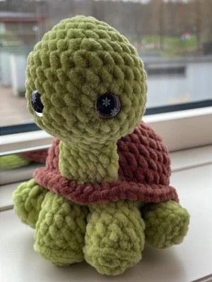 Virkad sittande sköldpaddsmjukis - Supergullig sittande handvirkad sköldpadda i mjukt grönt och brunt sammetsgarn. Sköldpaddan har stora svart/guldfärgade säkerhetsögon med vita detaljer och ett litet glittrigt hjärta på skalet. Perfekt som dekoration eller gosedjur till någon som gillar söta djur.