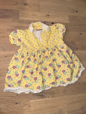 Gul blommig klänning från Kids Line - Gul blommig klänning från Kids Line Söt gul klänning med lila och gröna blommor från Kids Line. Klänningen har korta puffärmar, vit krage och spetskant nertill. Mönstret består av små vita blommor upptill och större blommor på kjolen. Perfekt för en färgglad look!
