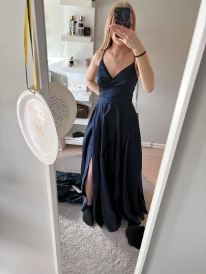 Marinblå långklänning med snörning - Elegant marinblå långklänning med v-ringad omlottringning och slits framtill. Klänningen har smala axelband som korsas och snörs i ryggen för en snygg detalj. Perfekt för bal eller festliga tillfällen.