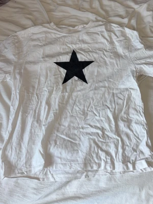 Vit t-shirt med svart stjärna Gina Tricot - Vit t-shirt från Gina Tricot med en stor svart stjärna tryckt på bröstet. Klassisk rund halsringning och korta ärmar. Tillverkad i mjuk bomull som känns skön mot huden. Perfekt för en enkel och cool look.